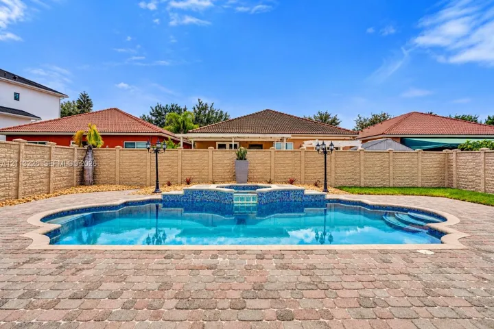 Custom-shaped in-ground swimming pool featuring a tiled spa with waterfall element, surrounded by paver decking and enclosed by a textured masonry privacy wall
