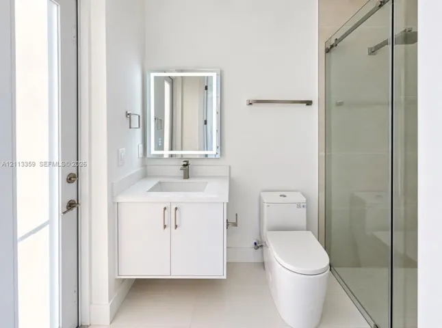 Floating vanity featuring a solid surface countertop, integrated sink, and brushed metal faucet, complemented by an illuminated rectangular mirror, a one-piece toilet, and a glass-enclosed shower with tile surround