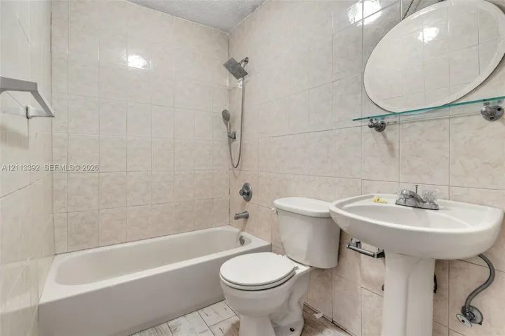 Full bathroom featuring tile-surround tub with hand-held showerhead, pedestal sink, oval wall-mounted mirror, and glass shelving