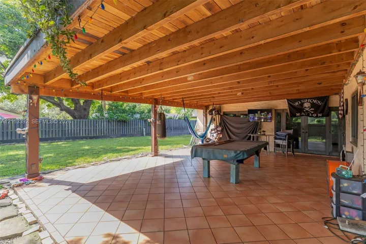 Covered patio featuring a timber-frame ceiling, tile flooring, and vertical support posts, overlooking a lawn enclosed by a privacy fence