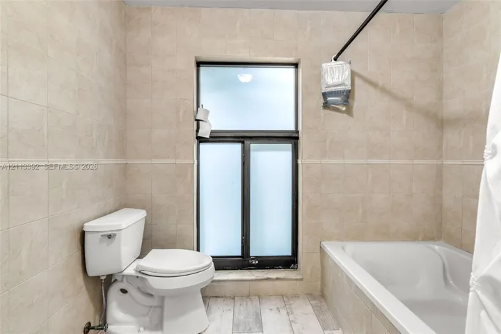 Bathroom featuring neutral tile wall cladding, wood-finish flooring, a white soaking tub, and a window with frosted glass panes