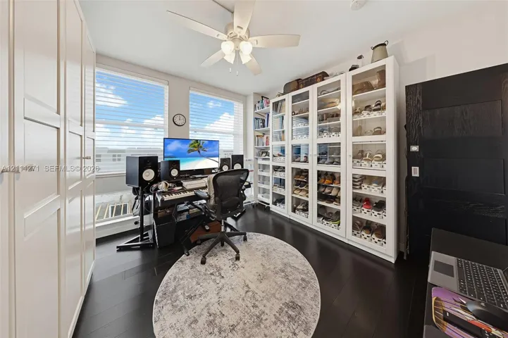 Room featuring dark-stained wood-finish flooring, floor-to-ceiling built-in white cabinetry with glass-front doors, tall windows with horizontal blinds, and a multi-blade ceiling fan with integrated lighting