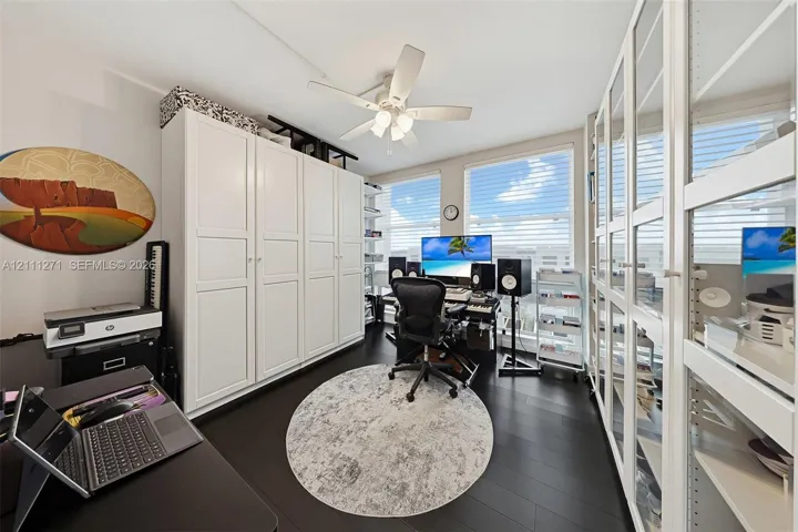 Custom built-in white cabinetry, expansive glass-paned storage units, dark wood-finish flooring, and a multi-blade ceiling fan with integrated lighting