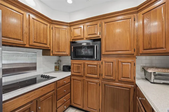 Kitchen featuring natural-finish wood cabinetry, a built-in microwave, a flush-mount electric cooktop, countertops, and a white subway tile backsplash