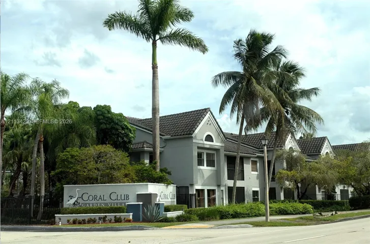 Multi-unit stucco building featuring a tile roof, screened-in balconies, mature palm trees, and professionally manicured landscaping