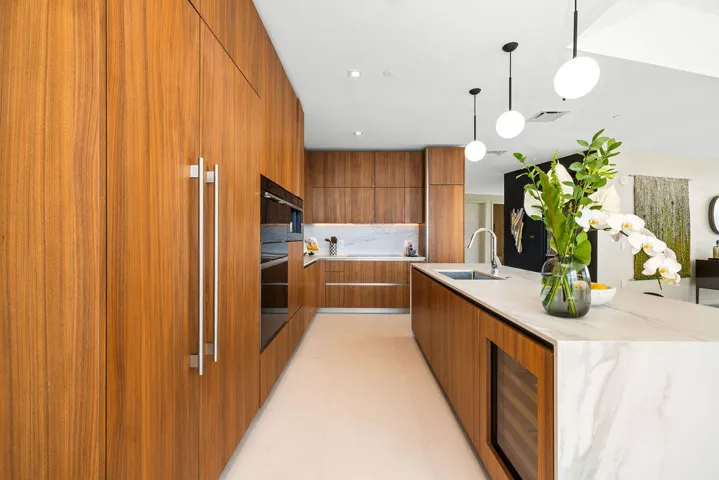 Modern kitchen featuring floor-to-ceiling wood-finish cabinetry, a waterfall-edge stone island, an integrated wine chiller, built-in wall oven, and pendant globe lighting