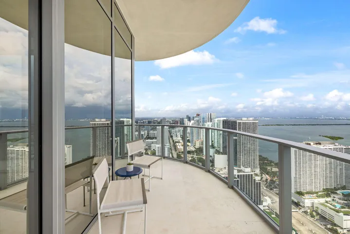 Curved private balcony featuring floor-to-ceiling glass wall, light-toned stone tile flooring, and a metal railing