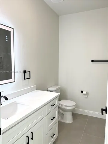 White vanity cabinet featuring a quartz countertop, integrated rectangular sink, matte black hardware, and an illuminated wall-mounted mirror