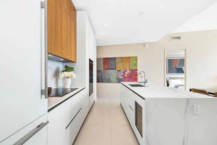 Kitchen featuring white flat-panel cabinetry, wood-finish upper cabinets, stone-surfaced waterfall island, built-in wall oven, and integrated wine cooler