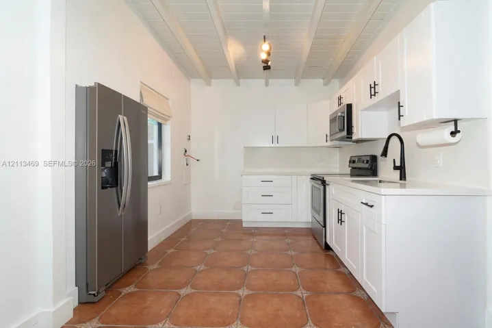 Kitchen featuring white shaker-style cabinetry, matte black hardware, stainless steel appliances, terracotta-toned tile flooring, and a beamed ceiling