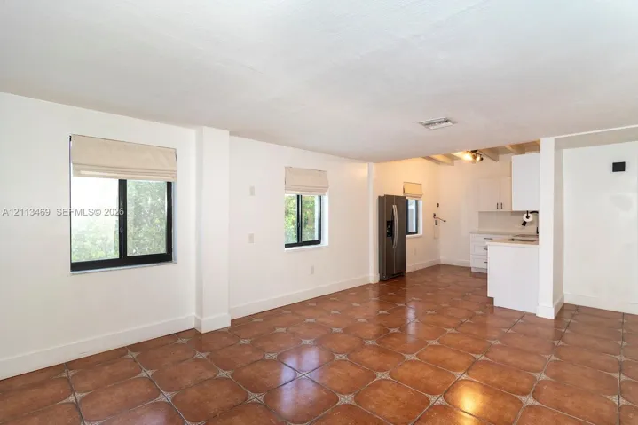 Open-concept interior featuring earth-tone ceramic tile flooring, neutral wall finishes, multi-pane windows, stainless steel refrigerator, and white cabinetry