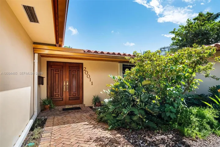 Stucco exterior entry featuring a herringbone-patterned brick walkway, double wood-paneled doors with metallic hardware, and a terracotta tile roof edge