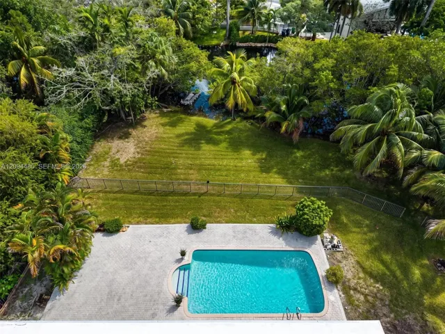 Outdoor living space featuring an in-ground swimming pool, concrete paver patio, metal perimeter fencing, manicured lawn, and mature tropical landscaping