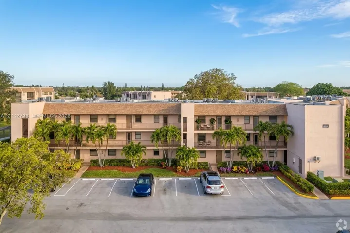 Multi-story exterior featuring a stucco facade, shingled roofing, paved surface parking, and mature palm tree landscaping