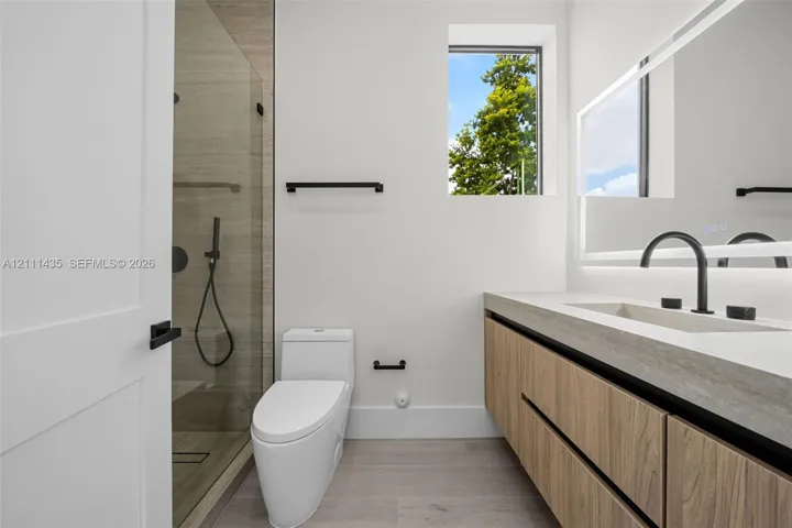 Floating vanity with wood-finish cabinetry, rectangular integrated sink, matte black hardware, glass-enclosed shower, and illuminated mirror