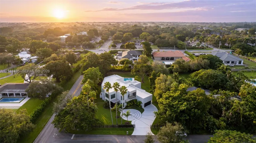 Aerial perspective of a multi-level contemporary residence featuring a flat roofline, circular concrete driveway, private rooftop pool, and mature perimeter landscaping