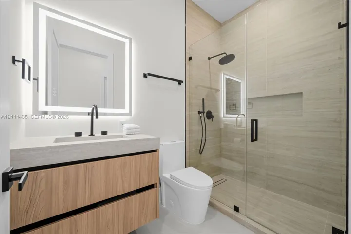 Floating wood-finish vanity with a rectangular sink, integrated LED-backlit mirror, walk-in shower with a glass enclosure and built-in recessed shelving, and matte black fixtures