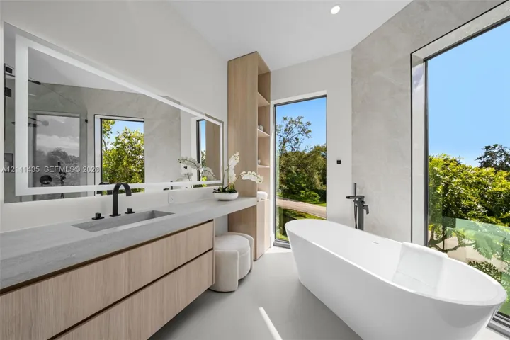 Floating wood-finish vanity with integrated sink, expansive LED-backlit mirror, freestanding soaking tub with floor-mounted faucet, and floor-to-ceiling windows