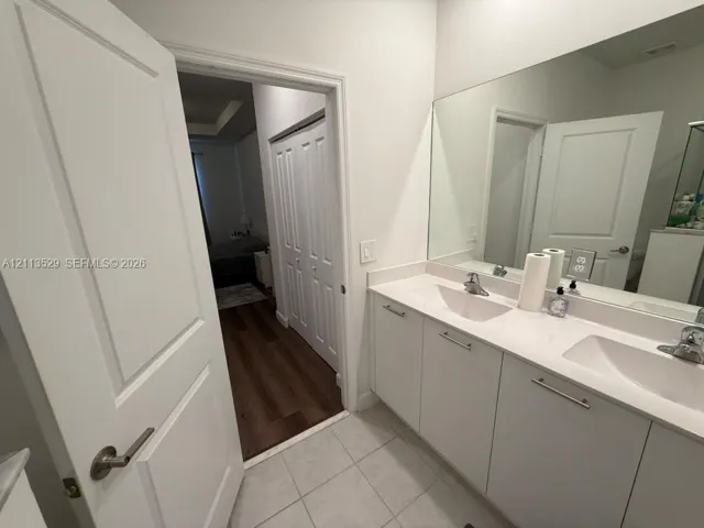 Bathroom featuring a dual-sink vanity with white cabinetry, solid-surface countertop, full-width mirror, and tile flooring