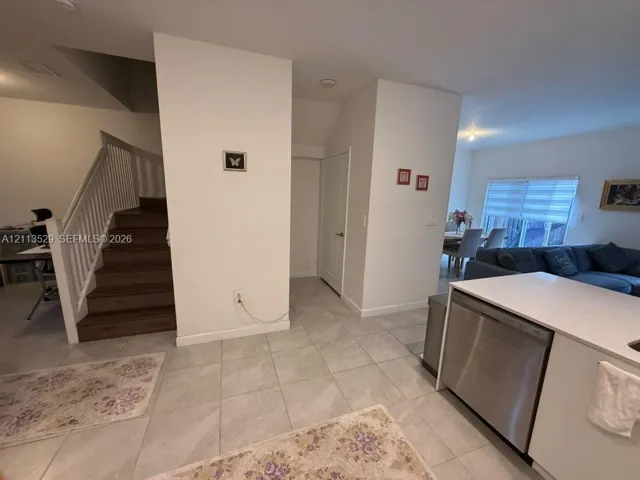 Open-concept interior featuring light-toned tile flooring, a staircase with wood-finish treads and white balusters, and a kitchen island with a stainless steel dishwasher and stone-look countertop