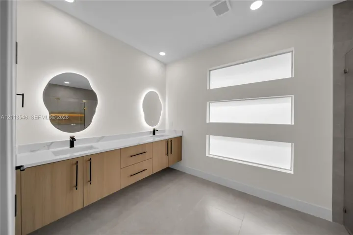 Floating vanity featuring wood-finish cabinetry, solid surface countertop with integrated dual sinks, matte black hardware, backlit organic-shaped mirrors, and horizontal transom windows