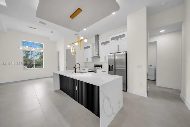 Kitchen featuring a central island with a stone-finish countertop and vertical slat detailing, white shaker-style cabinetry, stainless steel appliances, a modern multi-light pendant fixture, and large-format tile flooring