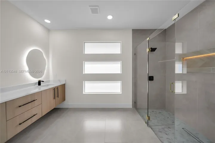 Floating wood-finish vanity with stone countertop, frameless glass walk-in shower with recessed tiled niche, and triple-stacked horizontal windows