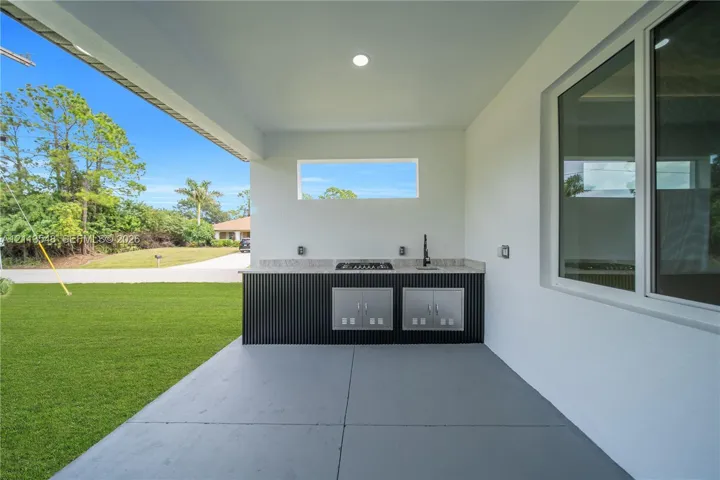 Covered outdoor kitchen featuring stainless steel built-in cabinetry, a stone-surfaced countertop, a cooktop, a sink with a gooseneck faucet, and concrete flooring