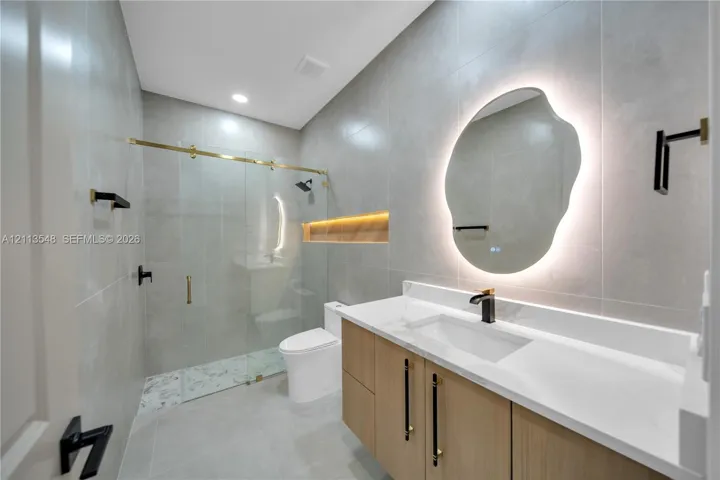 Bathroom featuring a floating wood-finish vanity with a stone countertop, backlit organic-shaped mirror, walk-in shower with sliding glass door and matte black fixtures, and recessed wall niche