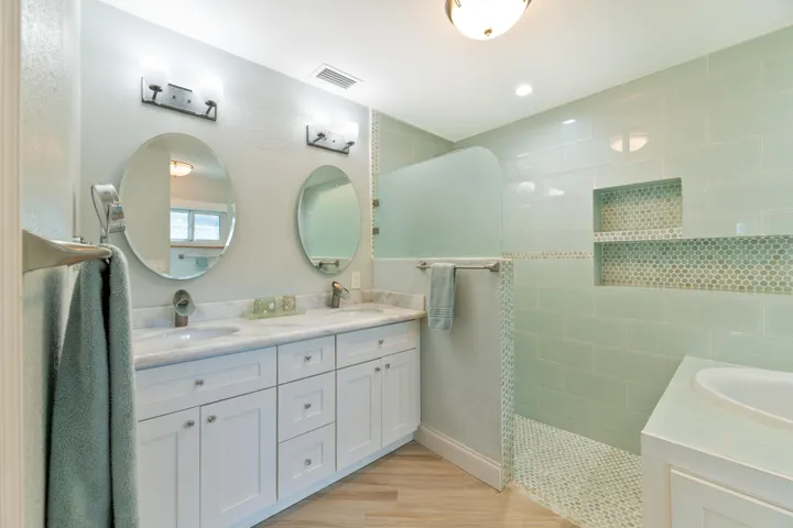 Dual-sink vanity with stone countertop, walk-in shower with tile surround and integrated shelving, wood-finish flooring, and dual oval wall mirrors