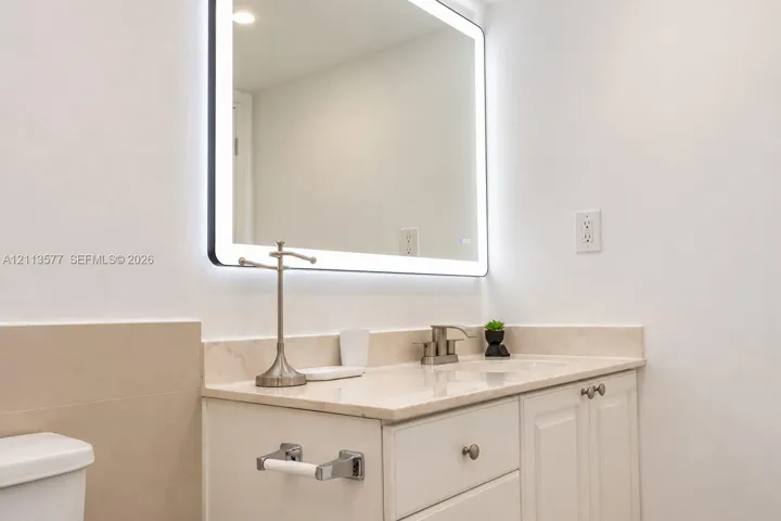 Rectangular LED-backlit mirror featuring rounded corners, paired with a white vanity cabinet, solid-surface countertop with integrated basin, and brushed-finish faucet