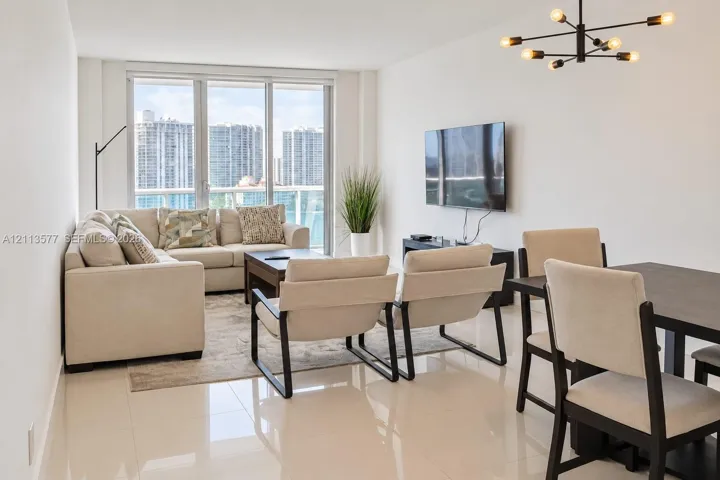 Living area featuring large-format tile flooring, floor-to-ceiling windows with a glass balcony railing, and a contemporary multi-arm chandelier
