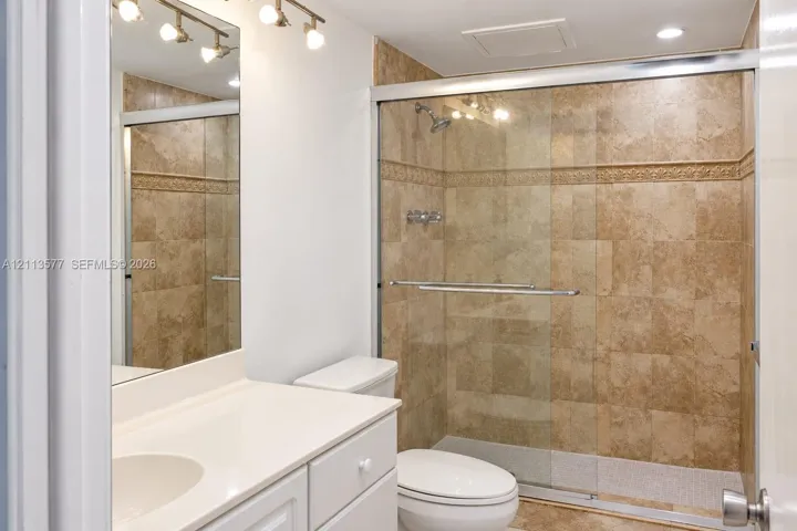 Bathroom featuring a vanity with a white countertop, an integrated sink, a full-length wall mirror, multi-light fixture, and a shower enclosure with floor-to-ceiling stone-finish tile surround and sliding glass doors