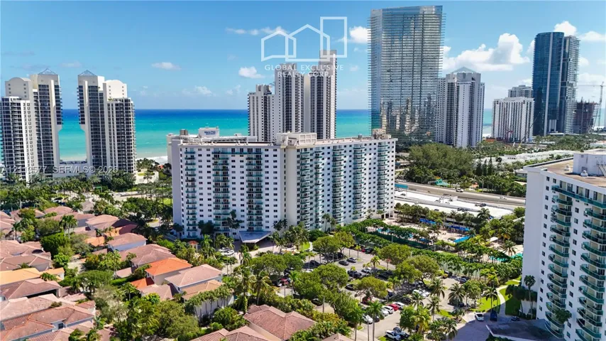 Aerial perspective featuring a multi-story residential building with private balconies, surrounded by mature palm trees, paved parking areas, and proximity to the coastline