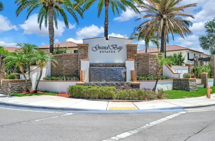 Community entrance featuring stacked-stone masonry, a built-in water feature, manicured landscaping, and mature palm trees