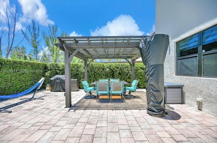 Outdoor patio area featuring a stone-paver surface, a fixed pergola, and a tall privacy hedge