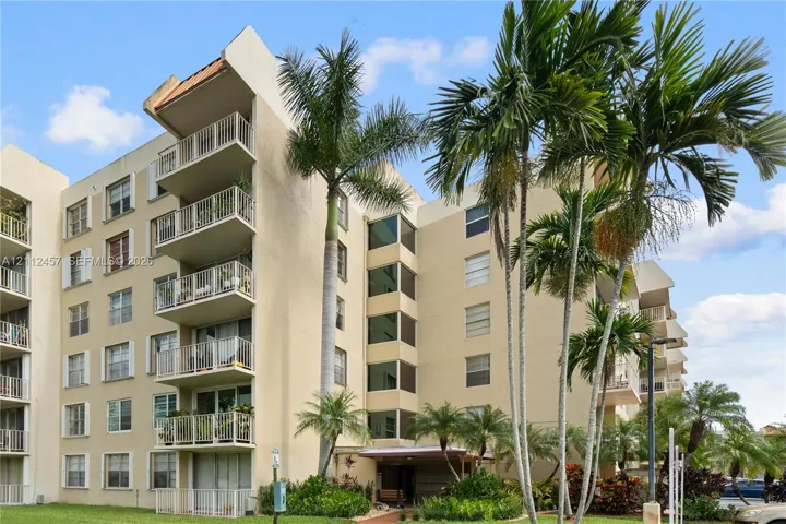Multi-story building exterior featuring stucco cladding, stacked private balconies with metal railings, and mature palm tree landscaping