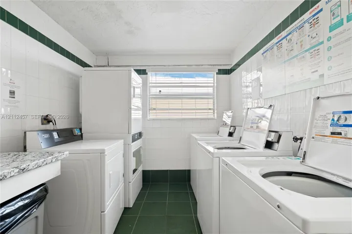 Laundry facility featuring green tile flooring, white wall-to-wall ceramic tiling with decorative accent trim, stone-finish countertop, and a window with horizontal blinds