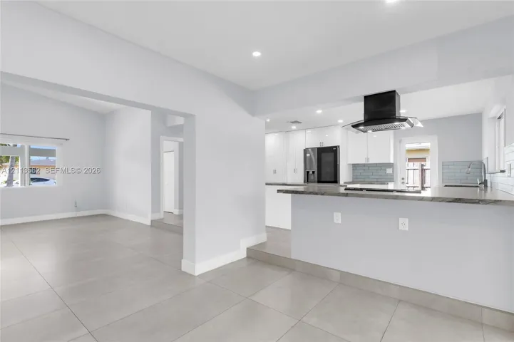 Open-concept kitchen featuring a breakfast bar, white cabinetry, dark stone-finish countertops, tile backsplash, and a black range hood