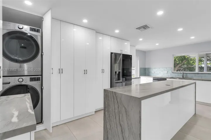 Kitchen featuring a waterfall-edge island with stone-finish countertop, high-gloss white cabinetry, integrated laundry station, and recessed lighting
