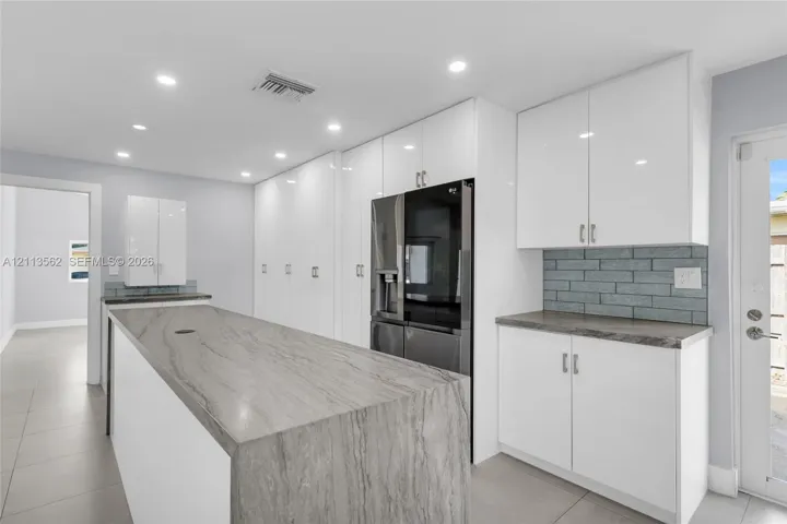 Kitchen featuring white flat-panel cabinetry, stone-surface waterfall island, grey brick-pattern backsplash, and tile flooring