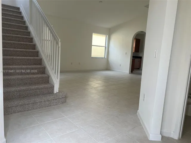 Open interior featuring neutral ceramic tile flooring, a carpeted staircase with white balusters and handrail, and an arched doorway