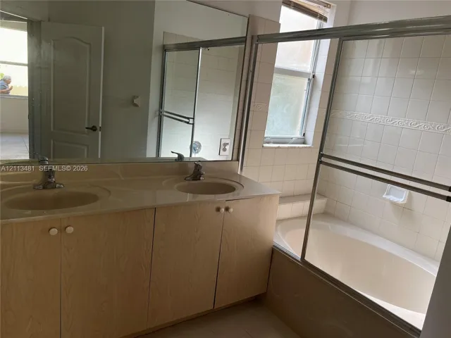 Bathroom featuring a dual-sink vanity with a solid-surface countertop, full-width wall mirror, soaking tub with a tiled surround, and a sliding glass shower enclosure
