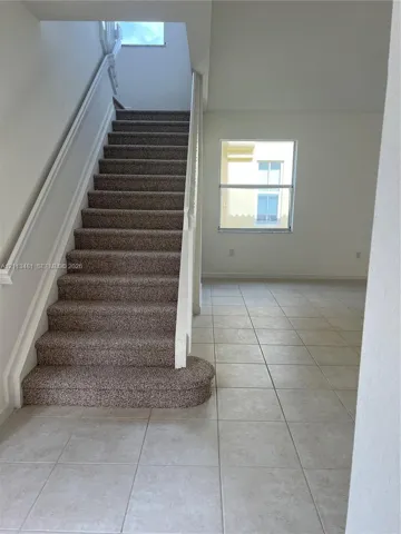 Staircase featuring carpeted treads, white handrails, and a curved bottom step, complemented by neutral tile flooring and a windowed wall