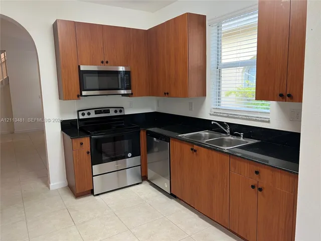 Kitchen featuring wood-finish cabinetry, dark solid-surface countertops, stainless steel appliances, double-basin sink, and ceramic tile flooring