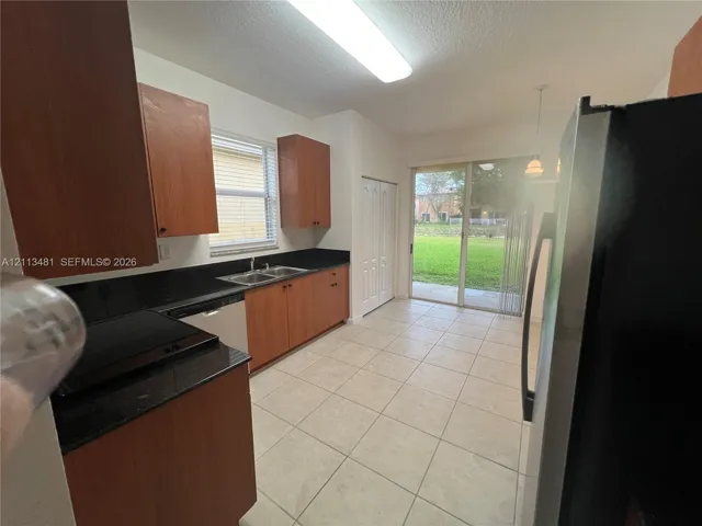Kitchen featuring wood-finish cabinetry, dark solid-surface countertops, double-basin stainless steel sink, tile flooring, and sliding glass door access to the rear exterior