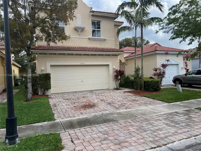 Two-story exterior featuring a stucco finish, mission-style clay tile roof, decorative corbel detail, and a paver driveway leading to an attached two-car garage