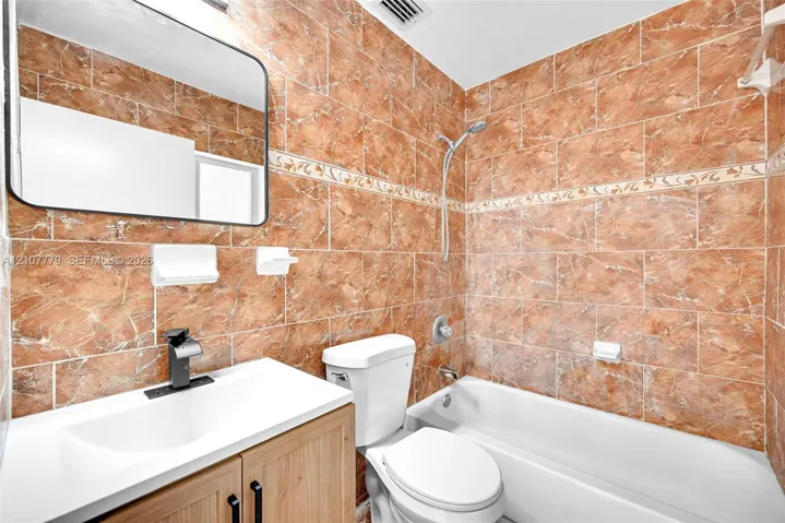 Bathroom featuring full-height ceramic tile walls with decorative inlay, a wood-finish vanity with an integrated countertop sink, matte black faucet hardware, and a frameless rectangular mirror