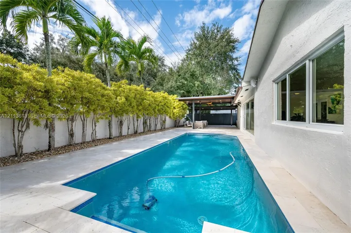 In-ground pool with stone-tiled decking, covered patio structure, and privacy wall surrounded by mature palm trees and greenery
