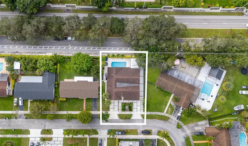 Aerial perspective of a property featuring a rectangular swimming pool, concrete paved driveway, structured landscaping, and a multi-section roofline
